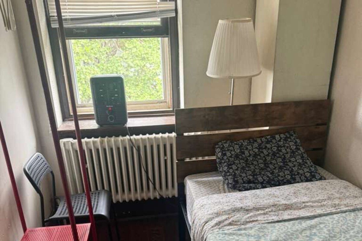 Una habitación con cama, una silla, un radiador y una ventana con persianas.