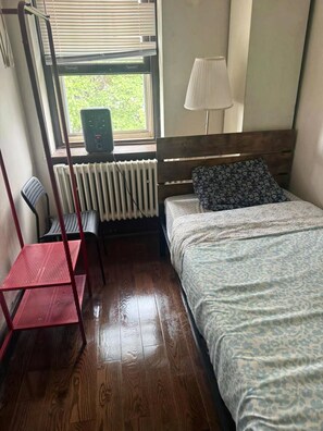 Una habitación con cama, una silla, un escritorio, una ventana con persianas, un radiador y una lámpara.