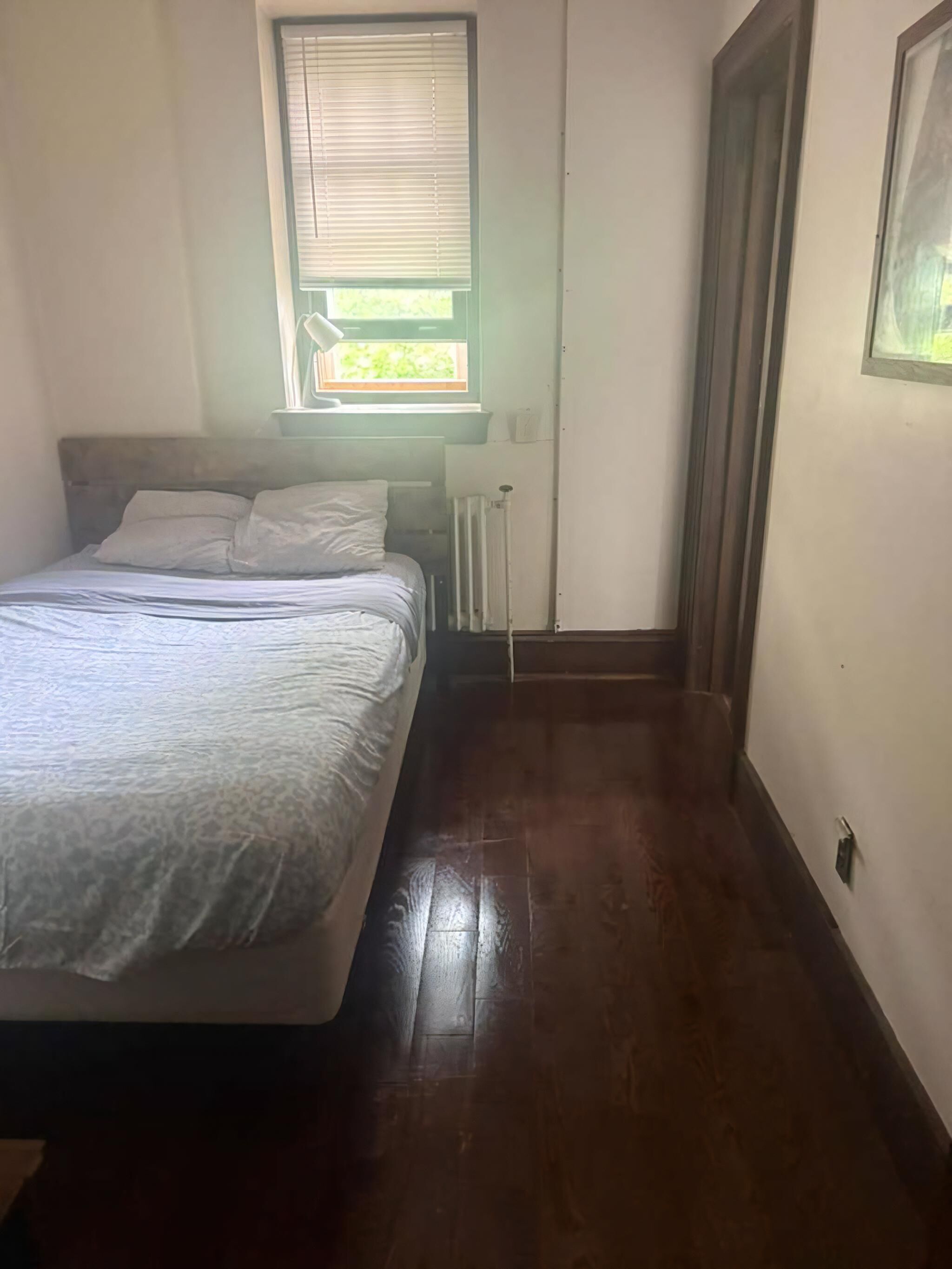A bedroom with a bed, a radiator, a window with blinds, and a door.