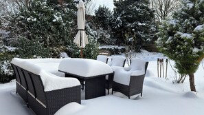 A snow-covered outdoor seating area with wicker furniture and a table, surrounded by snow-laden trees and a wooden fence.