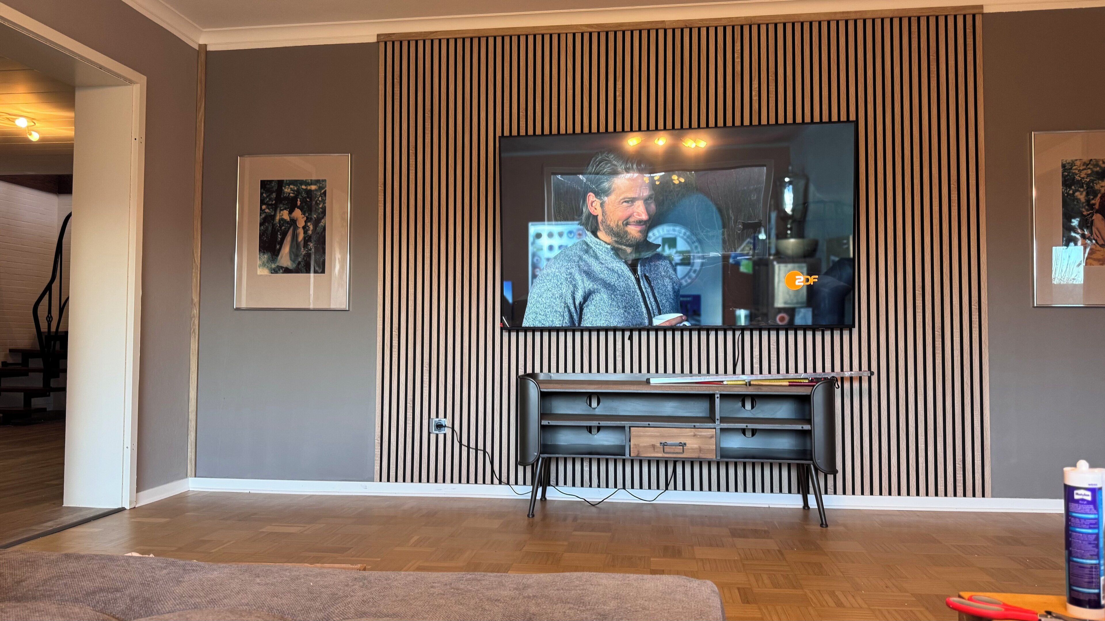 A living room with a flat-screen TV mounted on a wall with vertical wooden slats, a wooden entertainment unit below, and framed pictures on the walls.