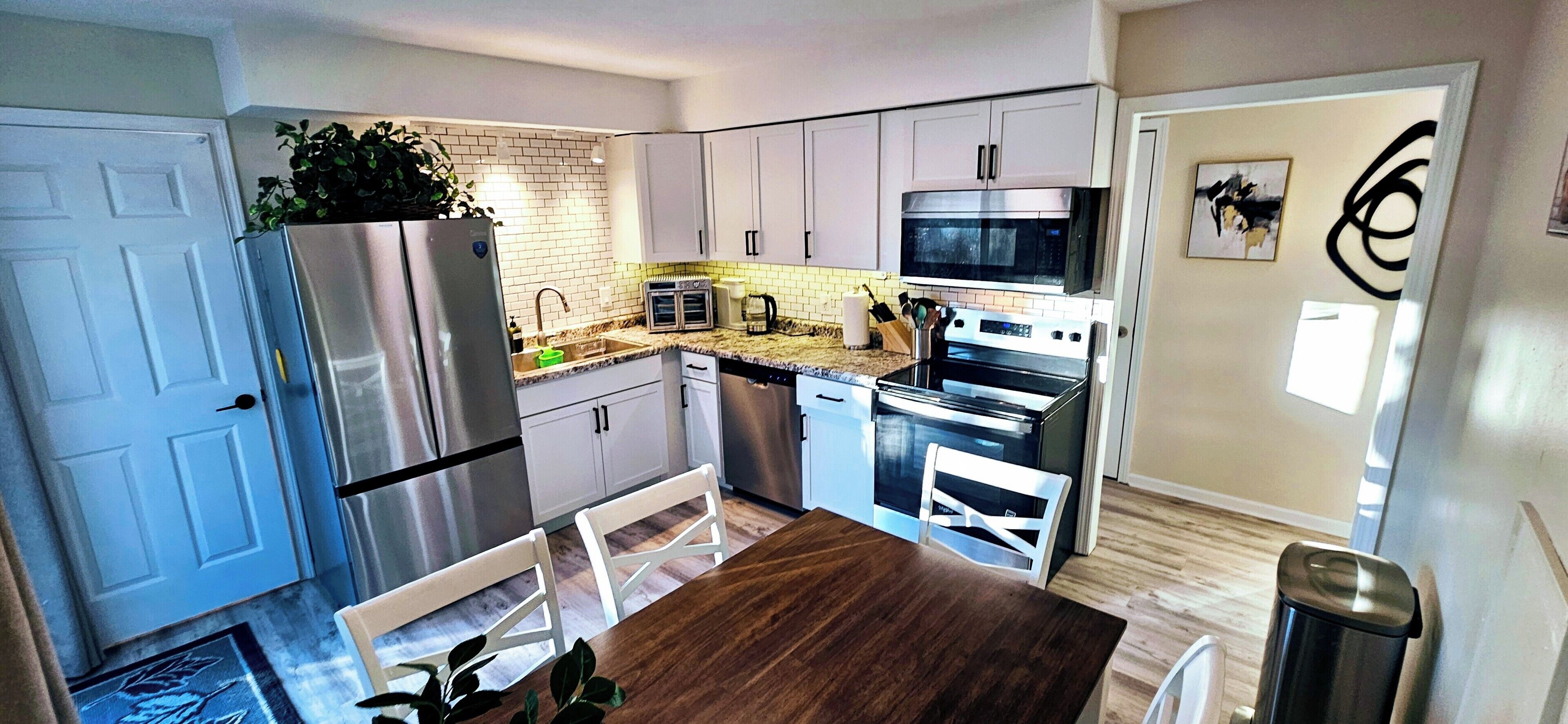 A modern kitchen with white cabinets, stainless steel appliances, a wooden dining table, and a potted plant.