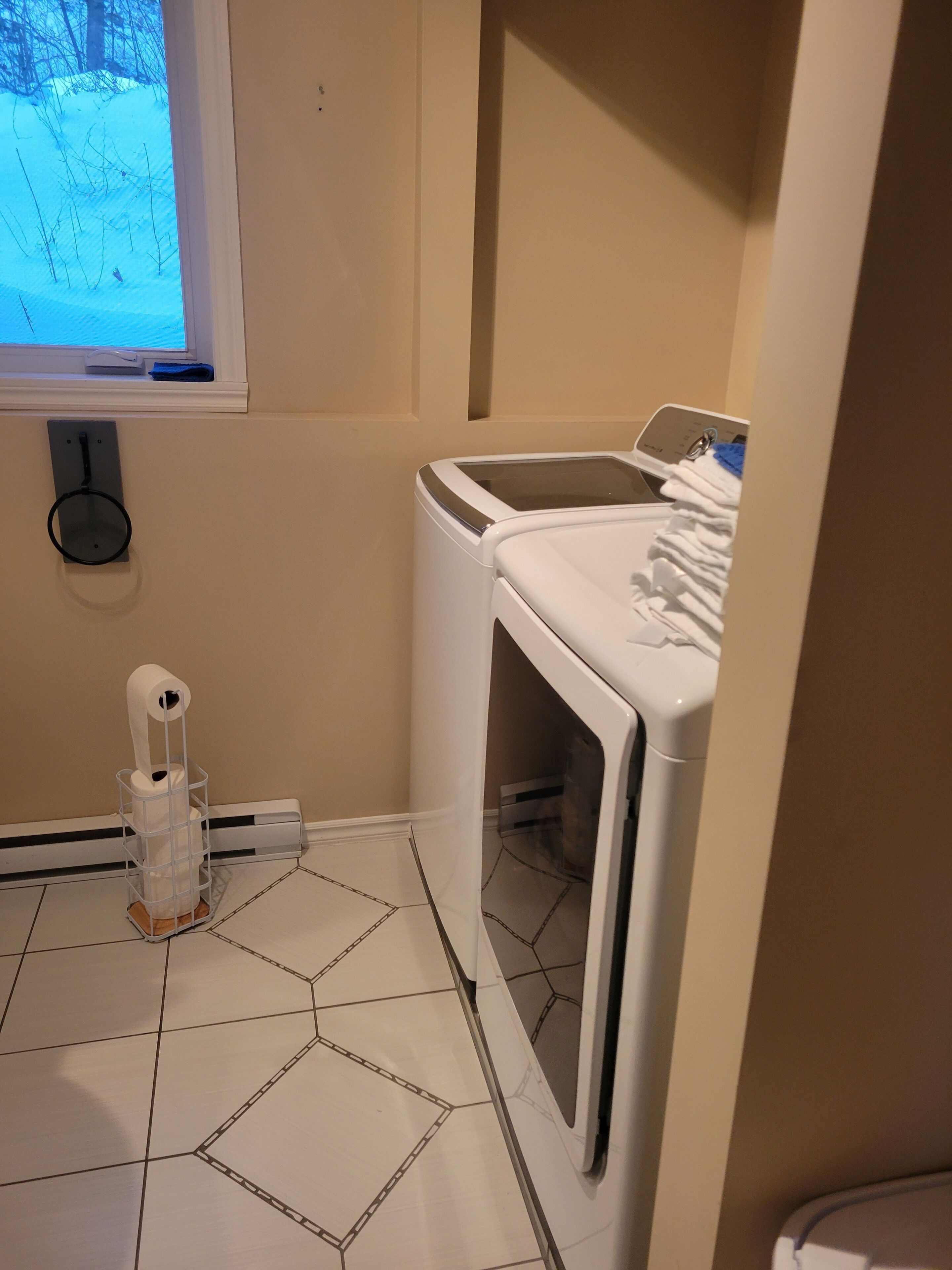 Una sala amb bugaderia que inclou una rentadora i una màquina d'assecar, un suport de paper higiènic, i una finestra amb vista a un paisatge nevàt.
