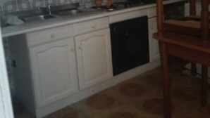 Cuina amb armaris blancs, campana extractora negra, respatller decorat i zona de dinar amb una cadira de fusta.