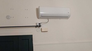 Seinään asennettava ilmastointilaite, jossa on virtajohto, seinäventtiili ja sen alapuolella ovi.