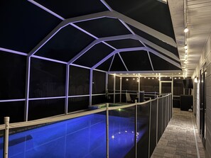 Ein überdachter Außenpool mit geometrischer Dachkonstruktion, einer Liegefläche am Pool und einem Weg zu einer Tür.
