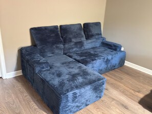 Un sofá modular con tela oscura y texturizada, ubicado frente a una pared color crema y suelo de madera.