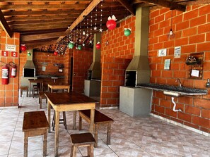 A brick-walled outdoor dining area with wooden tables and benches, a brick oven, and hanging decorations.