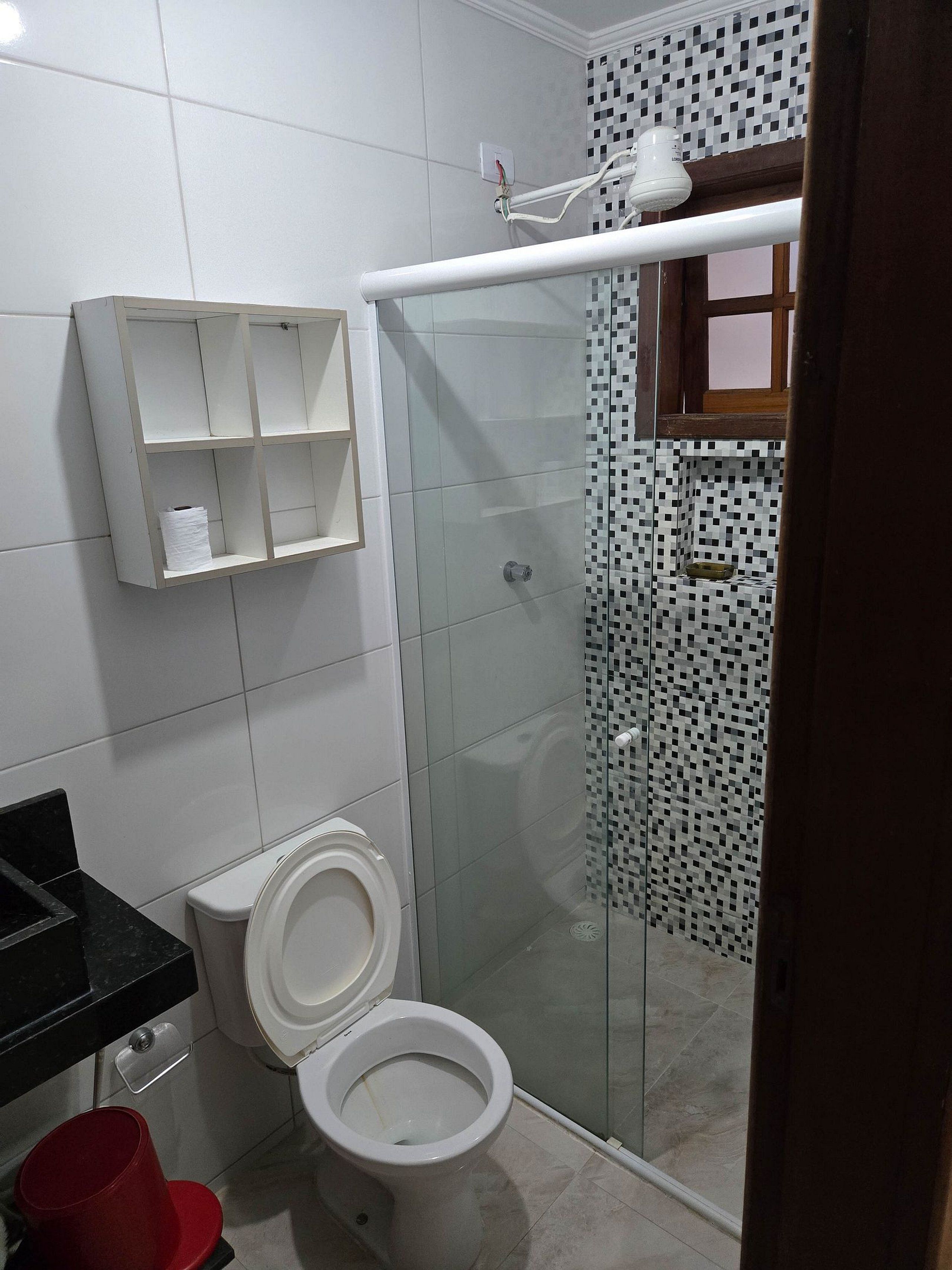 Un baño con inodoro, ducha con mampara de vidrio y pared de azulejos blancos.