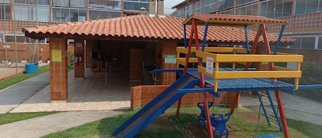 Um parquinho com escorrega e estrutura de escalada, rodeado por um edifício com janelas amplas e telhado cerâmico.
