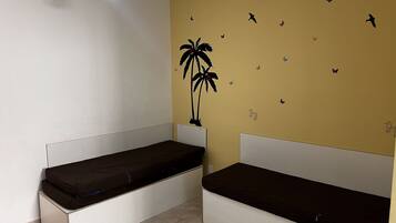Habitación con dos camas blancas y colchones oscuros, ventilador de techo y una pared de color amarillo decorada con calcomanías de pájaros y mariposas.