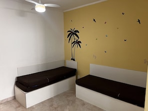 Une chambre avec deux lits blancs dotés de matelas foncés, un ventilateur de plafond et un mur de couleur jaune orné de motifs d’oiseaux et de papillons.