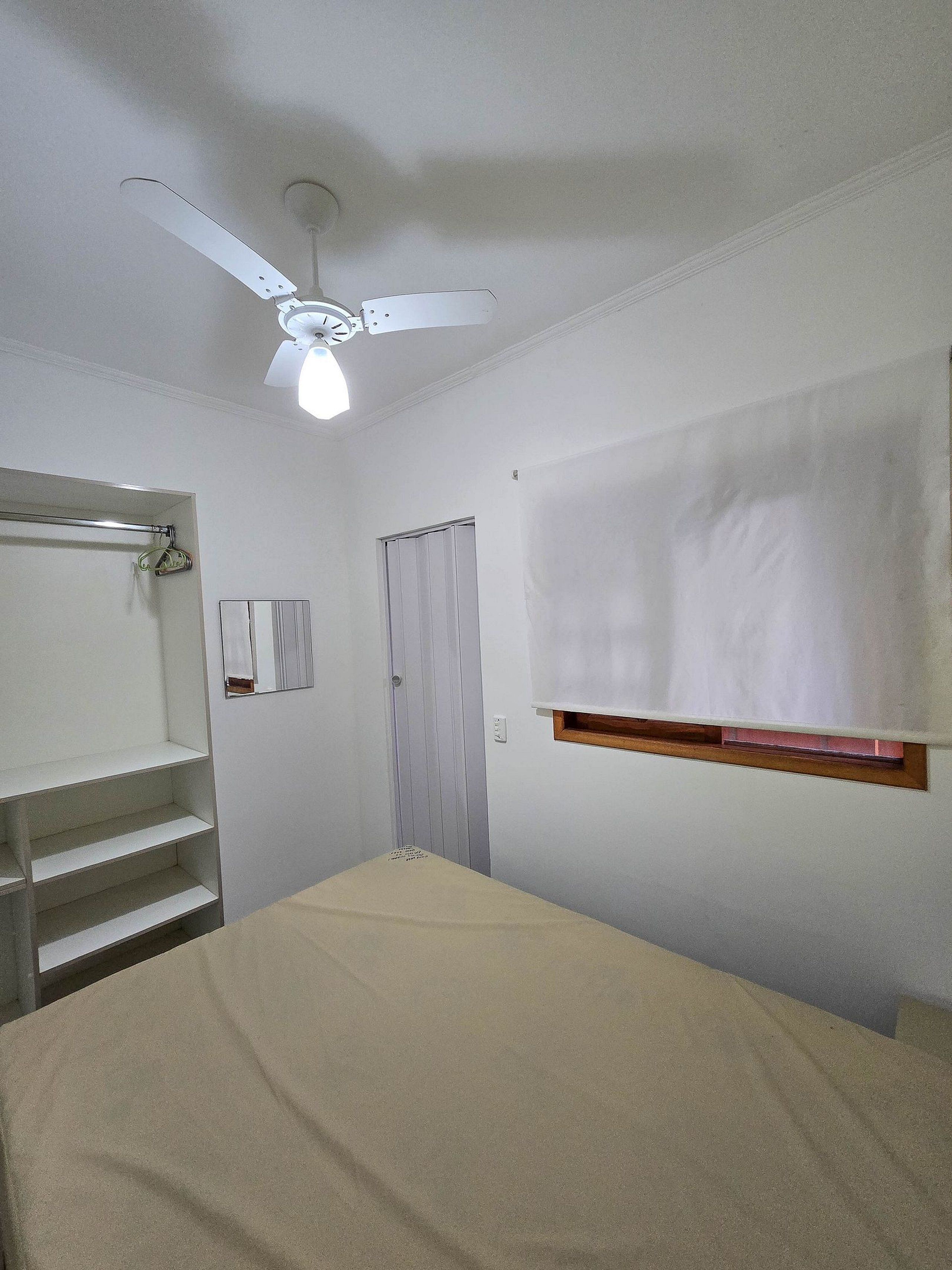 A bedroom with a bed, a ceiling fan, a window with blinds, a door, and a built-in shelf.