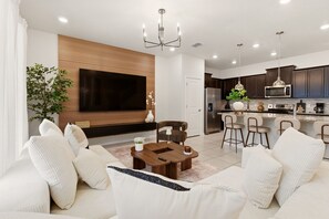 Ein modernes Wohnzimmer mit einem großen Flachbildfernseher, einer Wand mit Holzvertäfelung, einem Essbereich mit Holztisch und einer Küche mit dunklen Schränken und Edelstahlgeräten.