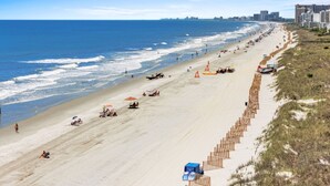 Een strand met een lange zandvlakte, parasols en ligbedden, met gebouwen op de achtergrond.