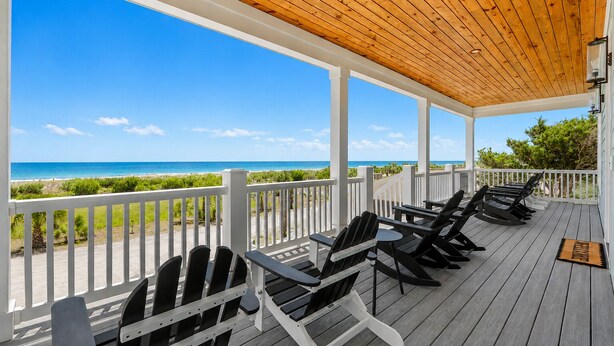 Een overdekt terras met houten vloer, witte leuningen en zwarte lounge fauteuils met uitzicht op het strand.
