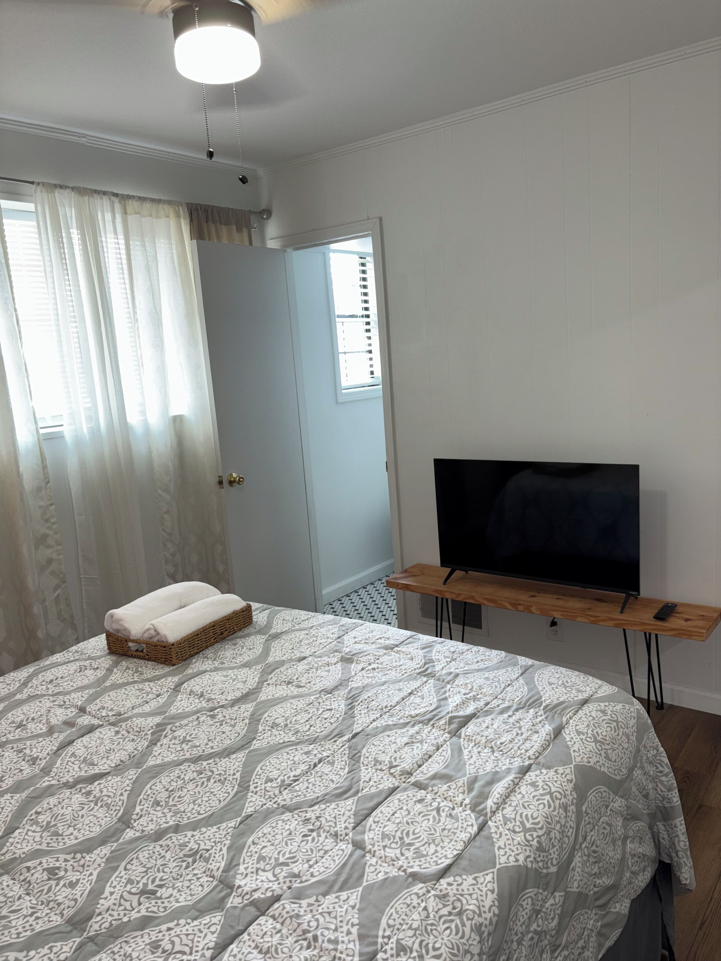 A bedroom with a bed, a TV on a wooden stand, a window with curtains, and a door.