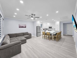 Ein moderner Wohnraum mit grauem Sofa, Esstisch mit Stühlen, Deckenventilator und Küchenzeile.