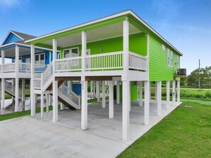 Ein zweistöckiges Haus mit grüner Fassade und weißen Geländern, das auf Stelzen steht, mit einer Veranda und einer Treppe, die zum oberen Stockwerk führt.