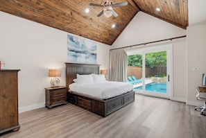 A bedroom with a wooden ceiling, a large bed, a nightstand, a lamp, and a view of a swimming pool through a sliding glass door. - Texas Pool Oasis, Luxury Amenities. Outdoor living. Fun for all. Dog friendly (Highland Village)