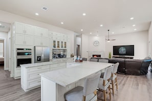 A modern kitchen with white cabinetry, a central island, and a dining area with a brown leather couch. - Texas Pool Oasis, Luxury Amenities. Outdoor living. Fun for all. Dog friendly (Highland Village)