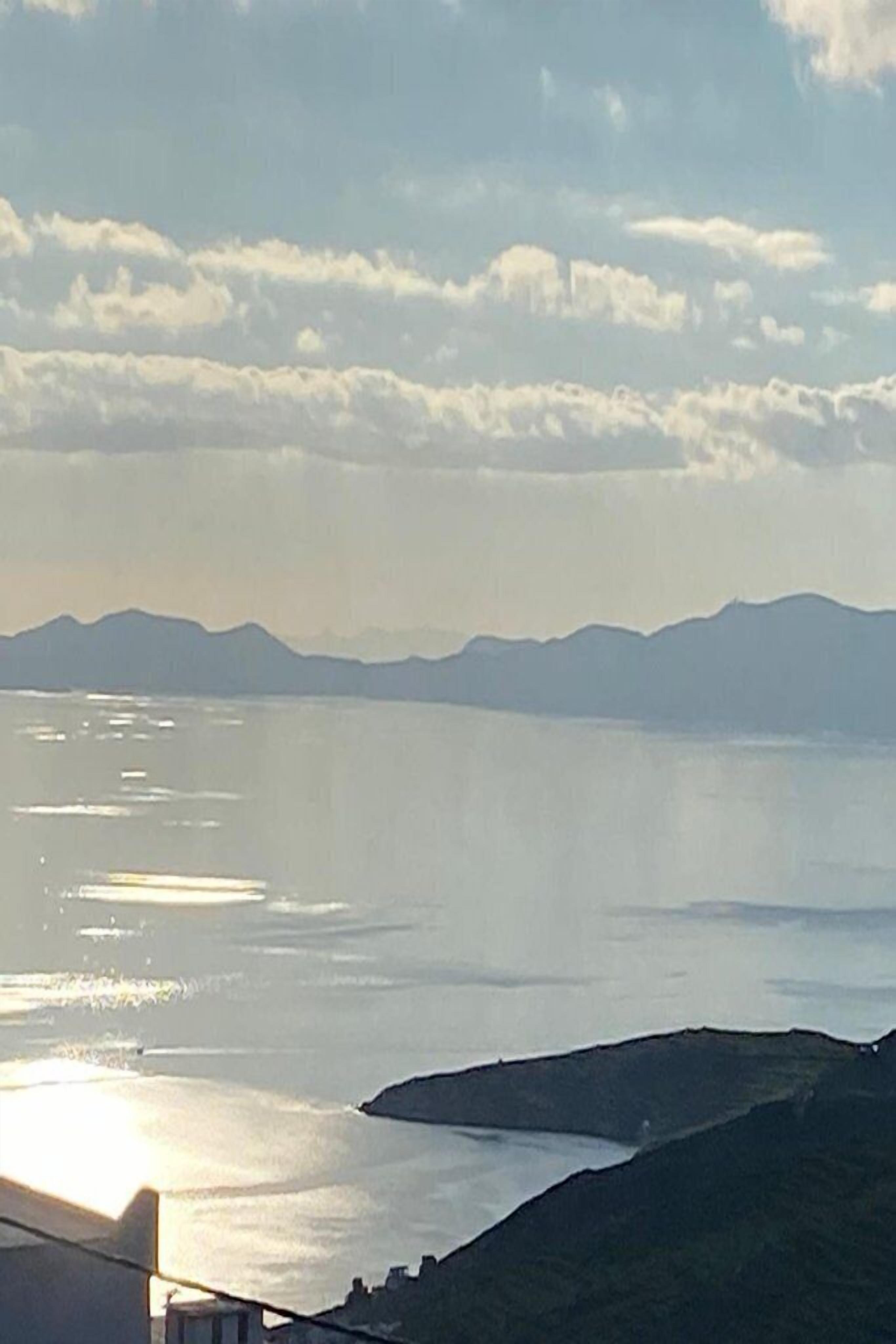 Eine Küstenlandschaft mit Bergen im Hintergrund, ein Gewässer, das Sonnenlicht reflektiert, und ein Gebäude mit einem geneigten Dach.