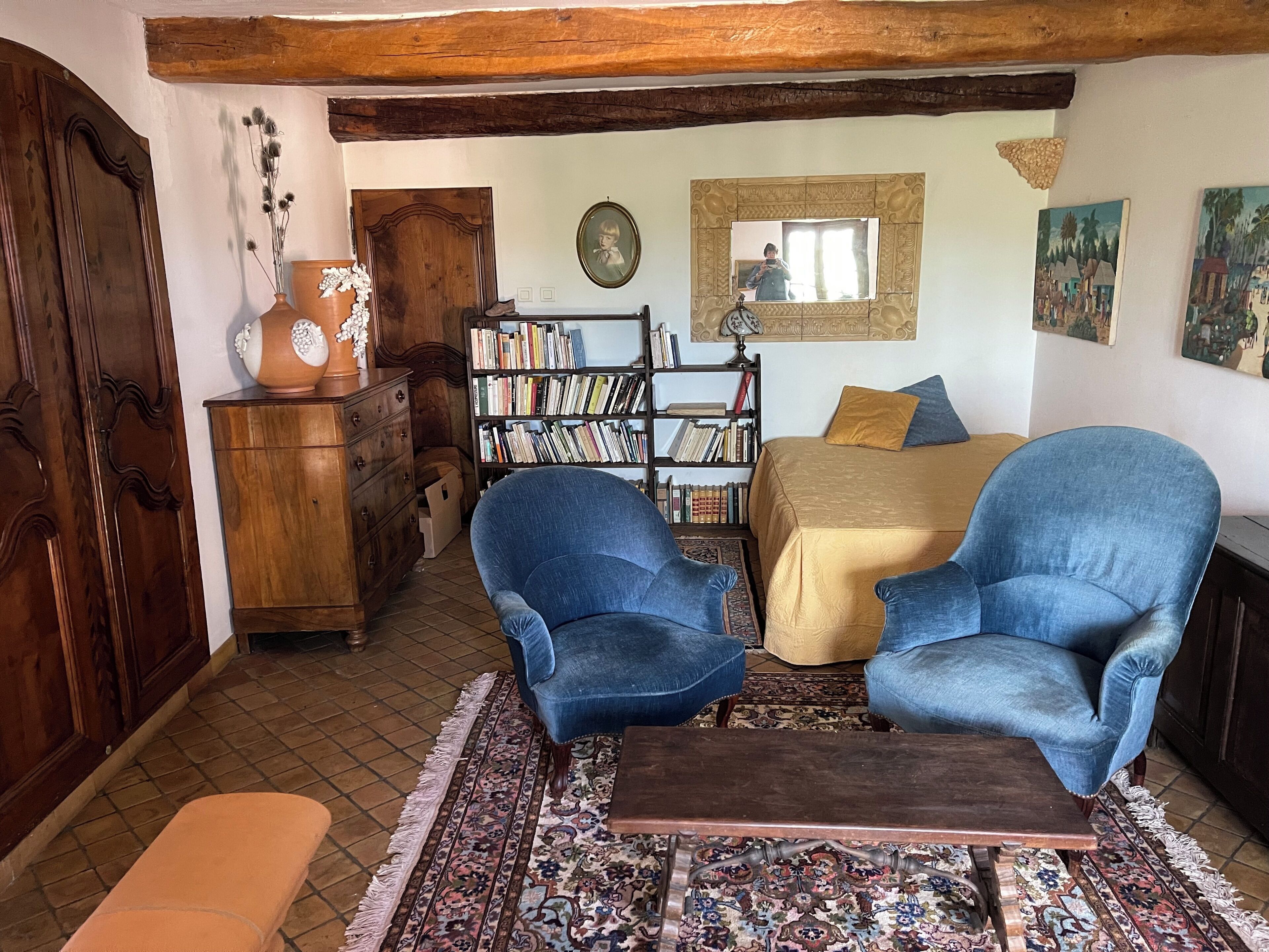 A cozy room with a bed, two blue armchairs, a wooden dresser, a bookshelf, and a patterned rug.
