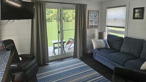 A room with a television, a sofa, a table, and a view of a green outdoor area.
