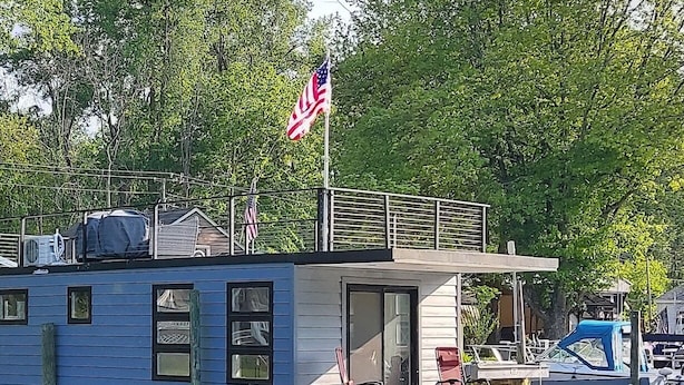 A houseboat with a flag, docked near a marina with boats and trees.