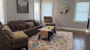 A living room with a brown sofa, patterned armchair, and ottoman, a coffee table with a decorative runner, a colorful rug, and a round mirror on the wall. - Modern Renovated Home near Six Flags & Naval Graduation Base - Sleeps 6 (Waukegan)