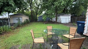 Un jardin avec une table en verre et des chaises en osier, entouré d’une clôture en treillis métallique et d’arbres.