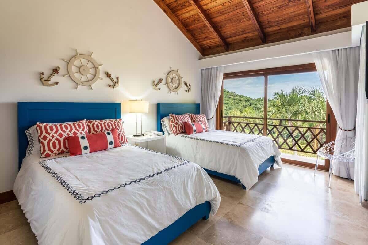 A room with two beds, a wooden ceiling, a balcony with a view of greenery, and decorative wall clocks.