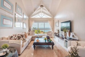 A spacious living room with a high ceiling, a large window offering a view of the sea, a comfortable sofa set, a coffee table with decorative items, and a television on a cabinet. - AEA- Samana Dominican Republic (Sanchez)