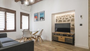 A living room with a sofa, a TV on a wooden stand, a stone wall, and a dining table with chairs. - Apartamentos Granadal. Bajo (Toledo)