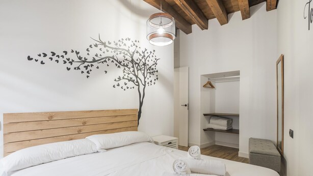 A bedroom with a wooden headboard, a wall-mounted tree decal, a bedside table, a mirror, and a walk-in closet. - Apartamentos Granadal. Bajo (Toledo)