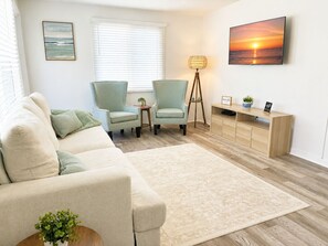 Ein Wohnzimmer mit einer Couch, zwei Sesseln, einem Couchtisch, einer Stehlampe, einem Fernseher und einem Bücherregal.
