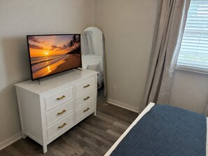 A bedroom with a TV on a white dresser, a bed with a blue blanket, and a window with blinds. - 10 Min to Gulf Coast Beaches - Pet Friendly - Fenced Yard (Seminole)