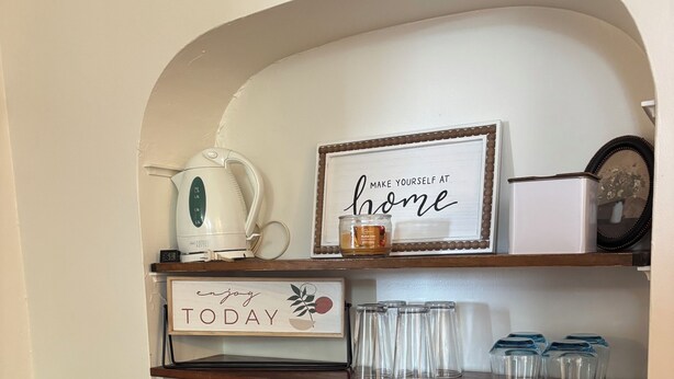 Una estantería de cocina con un hervidor blanco, una cafetera, un cartel enmarcado que dice 'Make Yourself at Home' y diversos utensilios de cocina.