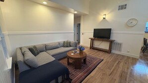 Un salon comprenant un canapé d’angle, une table basse ronde, un téléviseur à écran plat posé sur un meuble et un tapis à motifs.