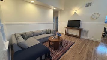 Un salon comprenant un canapé d’angle, une table basse ronde, un téléviseur à écran plat posé sur un meuble et un tapis à motifs.