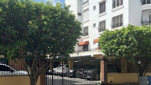 A gated residential area with a white building, trees, and parked cars. - Apartment (Santo Domingo)