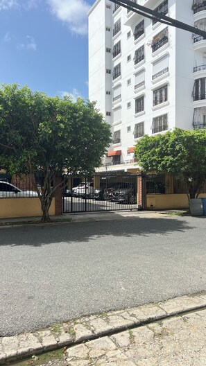 A gated residential area with a white building, trees, and parked cars. - Apartment (Santo Domingo)