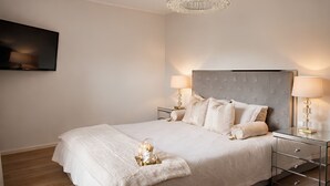 Une chambre à coucher avec un grand lit, deux tables de chevet avec des lampes, un téléviseur à écran plat fixé au mur et un lustre.