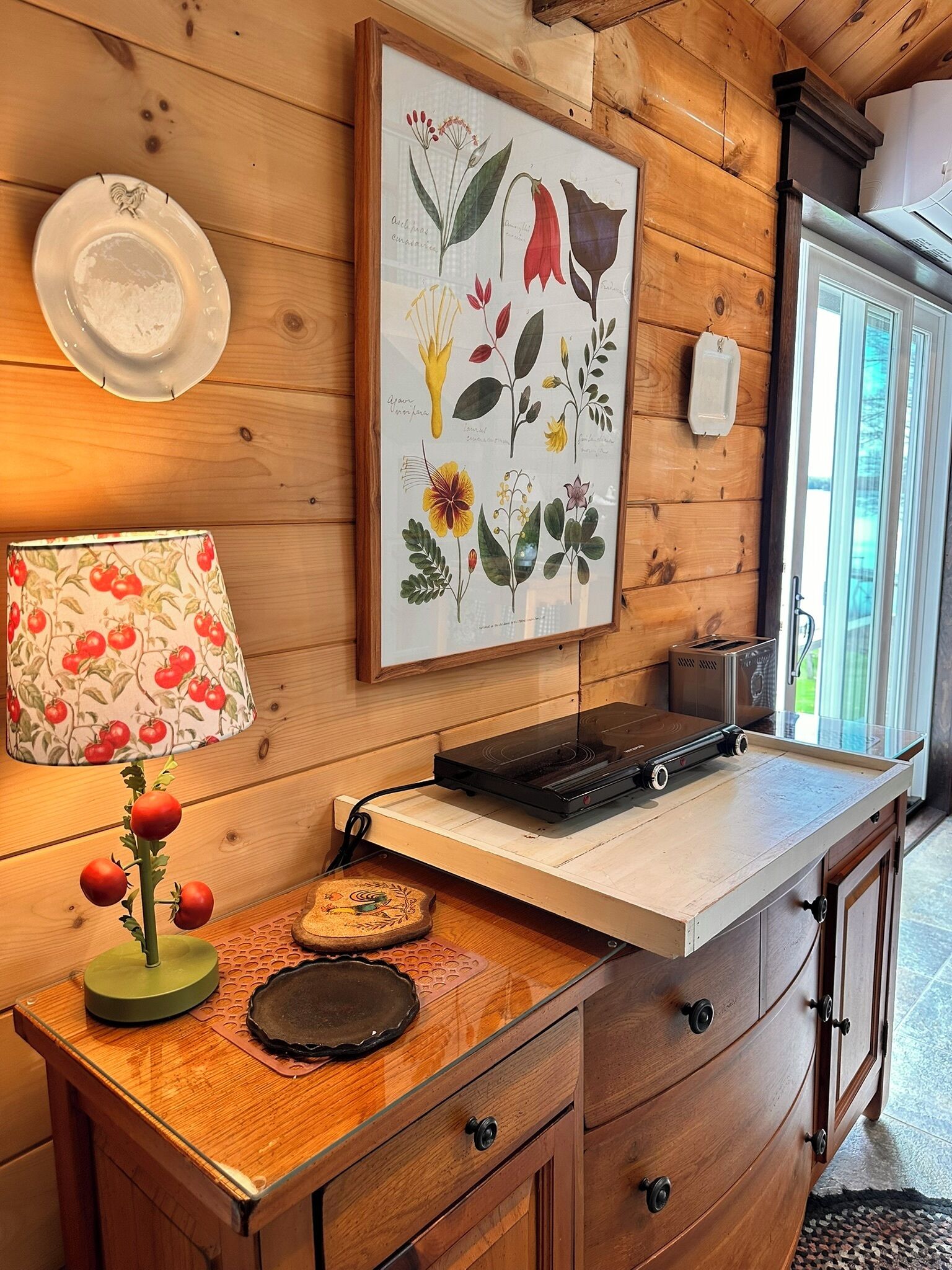 A wooden cabin interior with a kitchen counter, a framed botanical illustration, a lamp with cherry patterns, and a toaster.