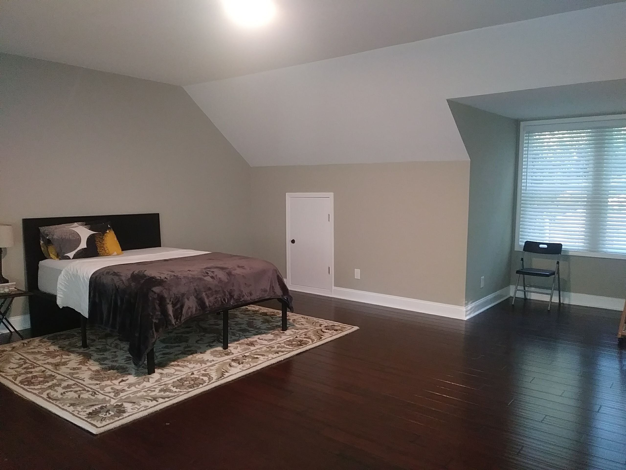Ein Schlafzimmer mit Bett, Nachttisch, Sessel, Fenster mit Jalousien und Tür.