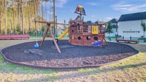 Um parquinho com um escorrega em formato de navio pirata, cercado por uma área circular de cobertura vegetal, com árvores e um edifício ao fundo.
