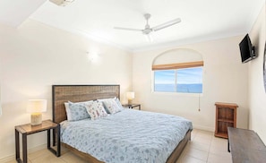 A bedroom with a bed, a ceiling fan, a nightstand with a lamp, a wooden nightstand, a TV mounted on the wall, and a window with a view. - Four, Villa Mar Colina (Yeppoon)