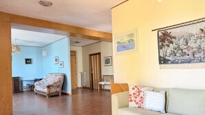 A living room with a sofa, a chair, and a dining table. There are framed pictures on the wall and a window with curtains. - C015 – Camerano – Three-story villa with garden (Camerano)