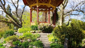 Un pabellón con estructura roja y amarilla, rodeado por un jardín con diversas plantas y una escalera de piedra que lleva a él.