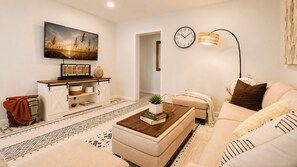A living room with a sofa, a coffee table with books, a TV, a wall clock, and a lamp. - 4BR Beach Getaway / 7 Min to Okaloosa Island / Fenced Yard / Sleeps 10 (Fort Walton Beach)